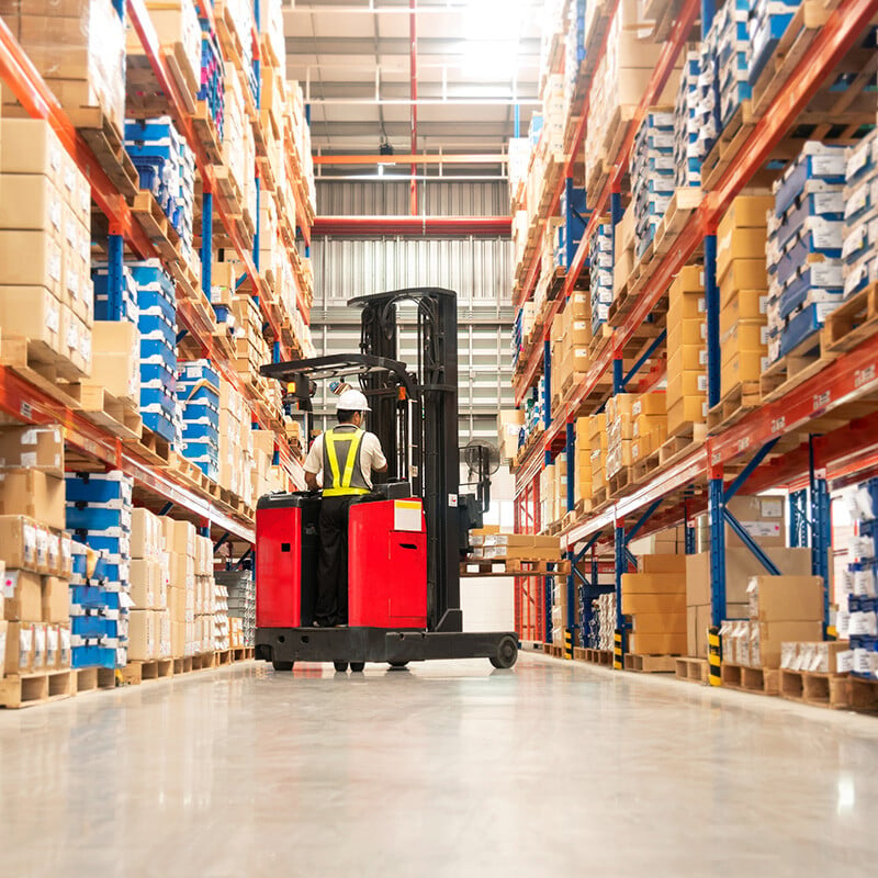 Worker in forklift-truck loading packed goods in huge distribution warehouse with high shelves