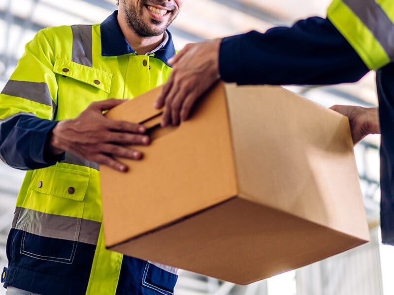 A warehouse worker handing a cardboard box to another warehouse worker